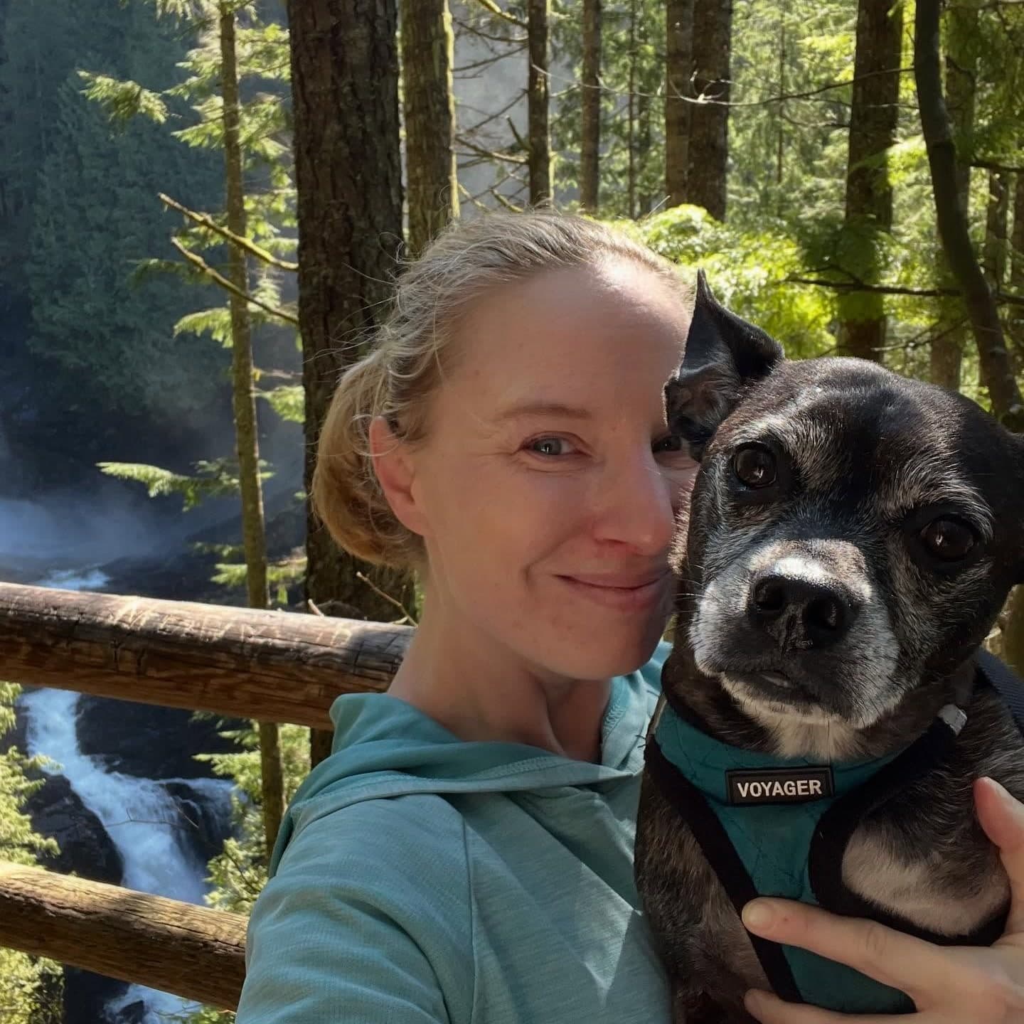 Julia with Diesel, their senior black brindle dog in a teal harness, at a forest waterfall overlook.